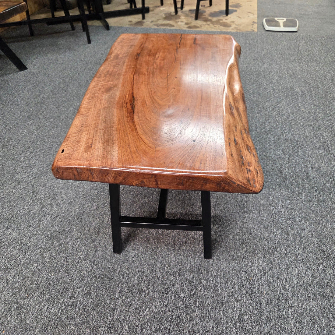 A live-edge wooden coffee table with a glossy medium-brown wood top and black metal legs, photographed in a showroom on a gray carpet.