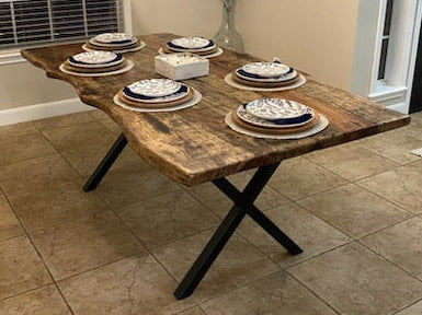 A rustic Pecan dining table with a live-edge natural top and black X-style legs, shown set with plates in a home dining room with beige tile floors and walls.