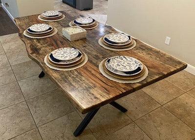 A rustic Pecan dining table with a live-edge natural top and black X-style legs, shown set with plates in a home dining room with beige tile floors and walls.