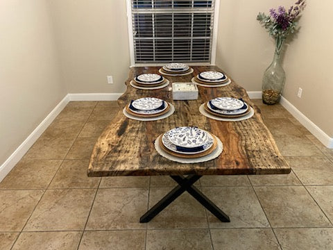 A rustic Pecan dining table with a live-edge natural top and black X-style legs, shown set with plates in a home dining room with beige tile floors and walls.