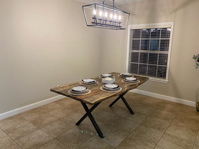 A rustic Pecan dining table with a live-edge natural top and black X-style legs, shown set with plates in a home dining room with beige tile floors and walls.