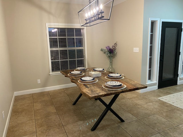A rustic Pecan dining table with a live-edge natural top and black X-style legs, shown set with plates in a home dining room with beige tile floors and walls.