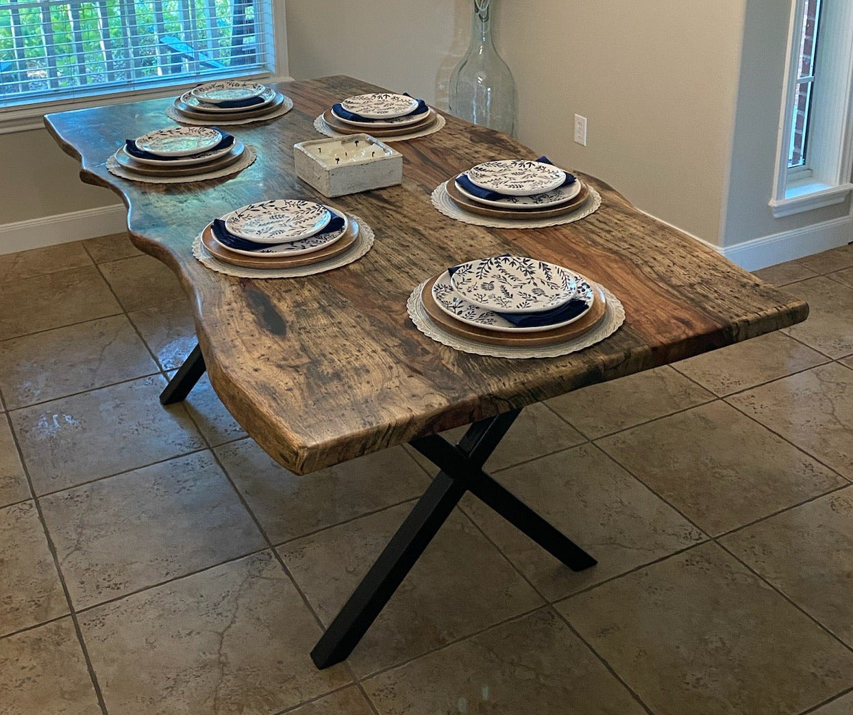 A rustic live-edge wood dining table with black X-style legs, set with plates and a vase of flowers in a dining room with beige walls and tiled flooring.