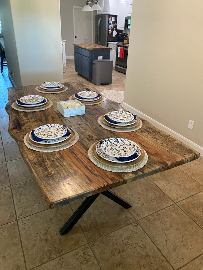 A rustic Pecan dining table with a live-edge natural top and black X-style legs, shown set with plates in a home dining room with beige tile floors and walls.