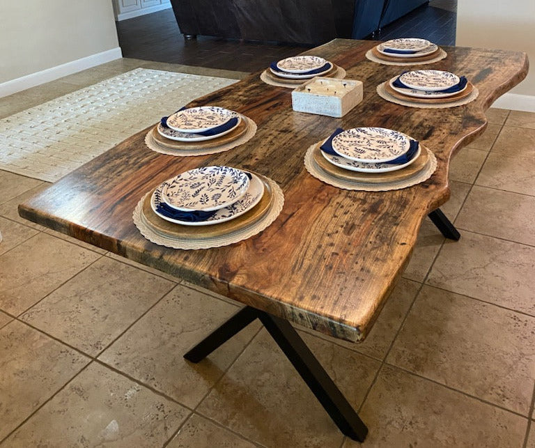 A rustic Pecan dining table with a live-edge natural top and black X-style legs, shown set with plates in a home dining room with beige tile floors and walls.