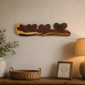 A rustic wooden wall-mounted decorative piece with three dark brown hearts on a natural wood base, displayed above a wooden console table with a vase, basket, framed sign, and lamp in a warm, styled interior.