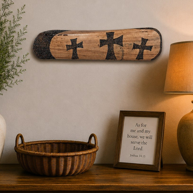 A rustic wooden wall plaque featuring three black crosses, styled on a wooden console table with a ceramic vase, basket, framed scripture print, and a lit table lamp in a warm, home interior setting.