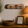 A rustic wooden wall plaque featuring three black crosses, styled on a wooden console table with a ceramic vase, basket, framed scripture print, and a lit table lamp in a warm, home interior setting.