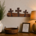 A rustic wooden wall decor piece featuring three crosses on a horizontal plank, styled on a beige wall above a wooden console table with a lamp, vase, basket, and framed scripture.