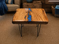 A rustic live-edge wooden coffee table with a glossy finish and a vibrant blue resin river running down the center, supported by black hairpin legs in a cozy living room setting.