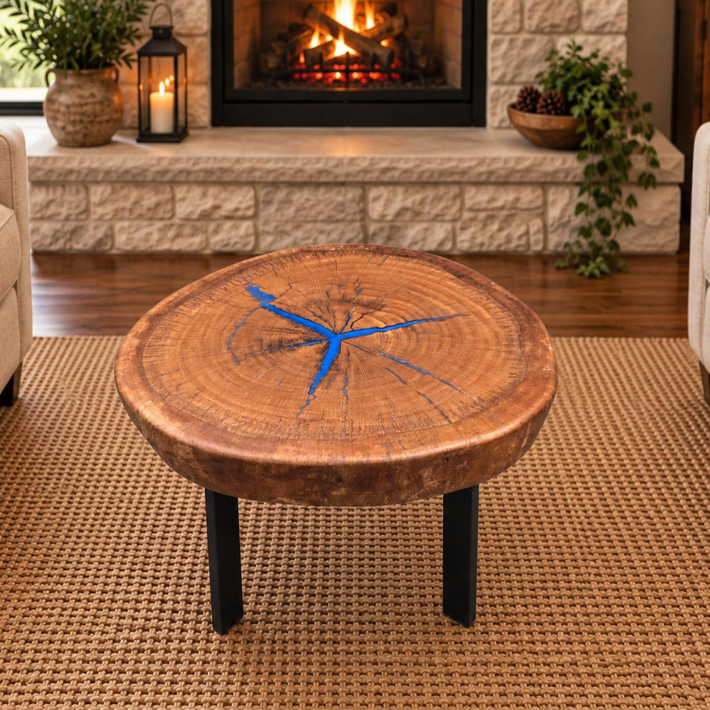 Round pecan coffee table with blue epoxy-filled cracks and black metal legs staged between two chairs in front of a stone fireplace.