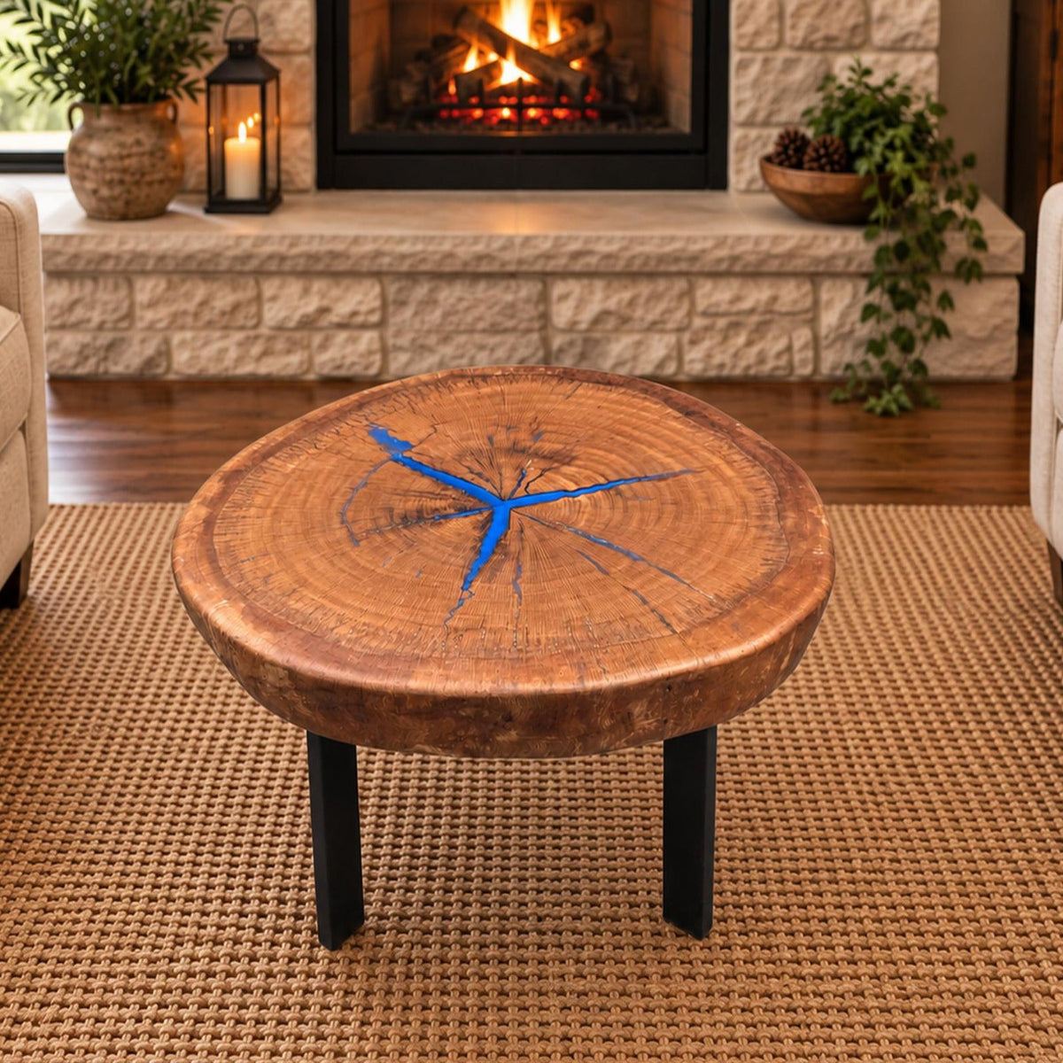 Round pecan coffee table with blue epoxy-filled cracks and black metal legs staged between two chairs in front of a stone fireplace.