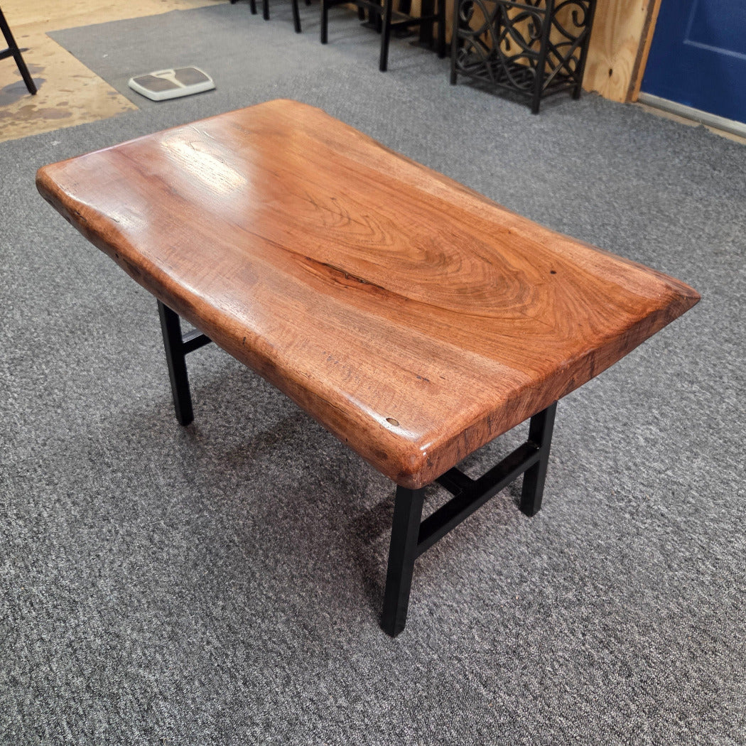 A live-edge wooden coffee table with a glossy warm brown finish and black metal legs, shown on a gray carpeted floor in what appears to be a showroom or warehouse.