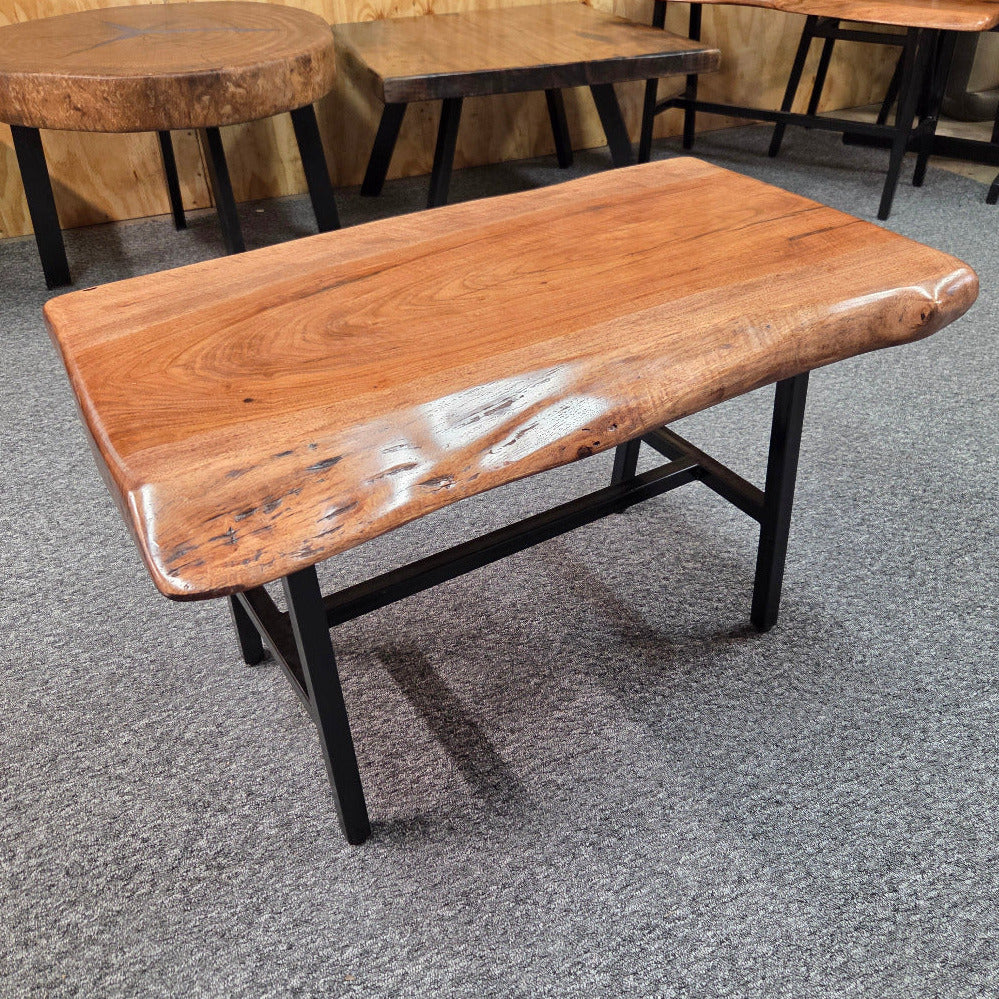 A live-edge wooden coffee table with a glossy brown wood top and black metal legs, shown on a gray carpeted floor in a furniture showroom.