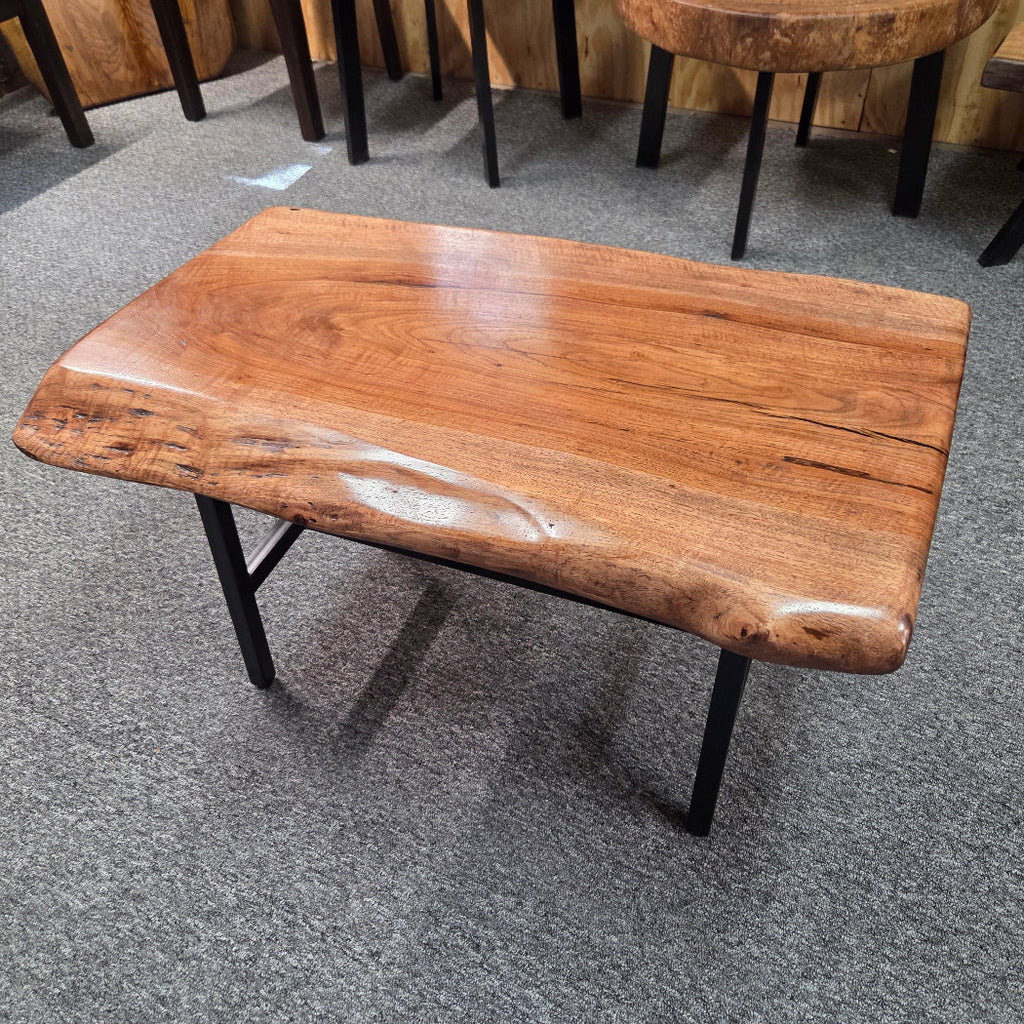 A rectangular live-edge wooden coffee table with a glossy warm brown finish and black metal legs, shown on a gray carpeted floor in a furniture showroom.