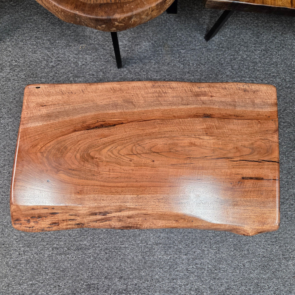 A rectangular live-edge wooden board with a glossy finish, showcasing rich warm brown wood grain, photographed from a top-down angle on a gray carpeted floor.