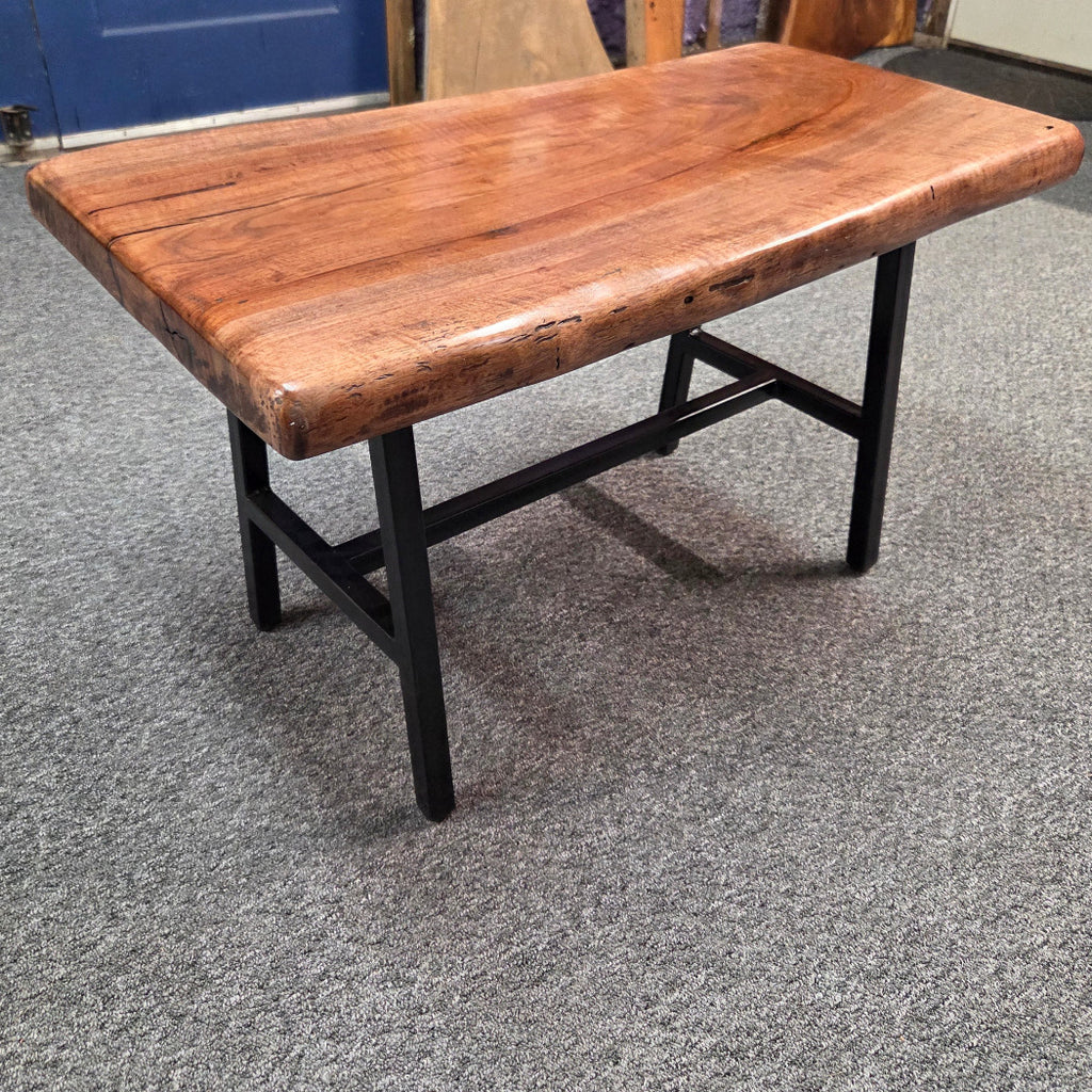 A rustic rectangular coffee table with a thick, polished live-edge wood top in warm brown tones and a black metal frame, shown on a gray carpeted floor.
