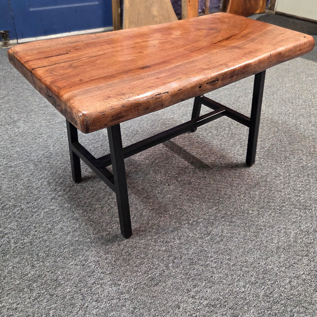A rustic rectangular coffee table with a thick, polished live-edge wood top in warm brown tones and a black metal frame, shown on a gray carpeted floor.
