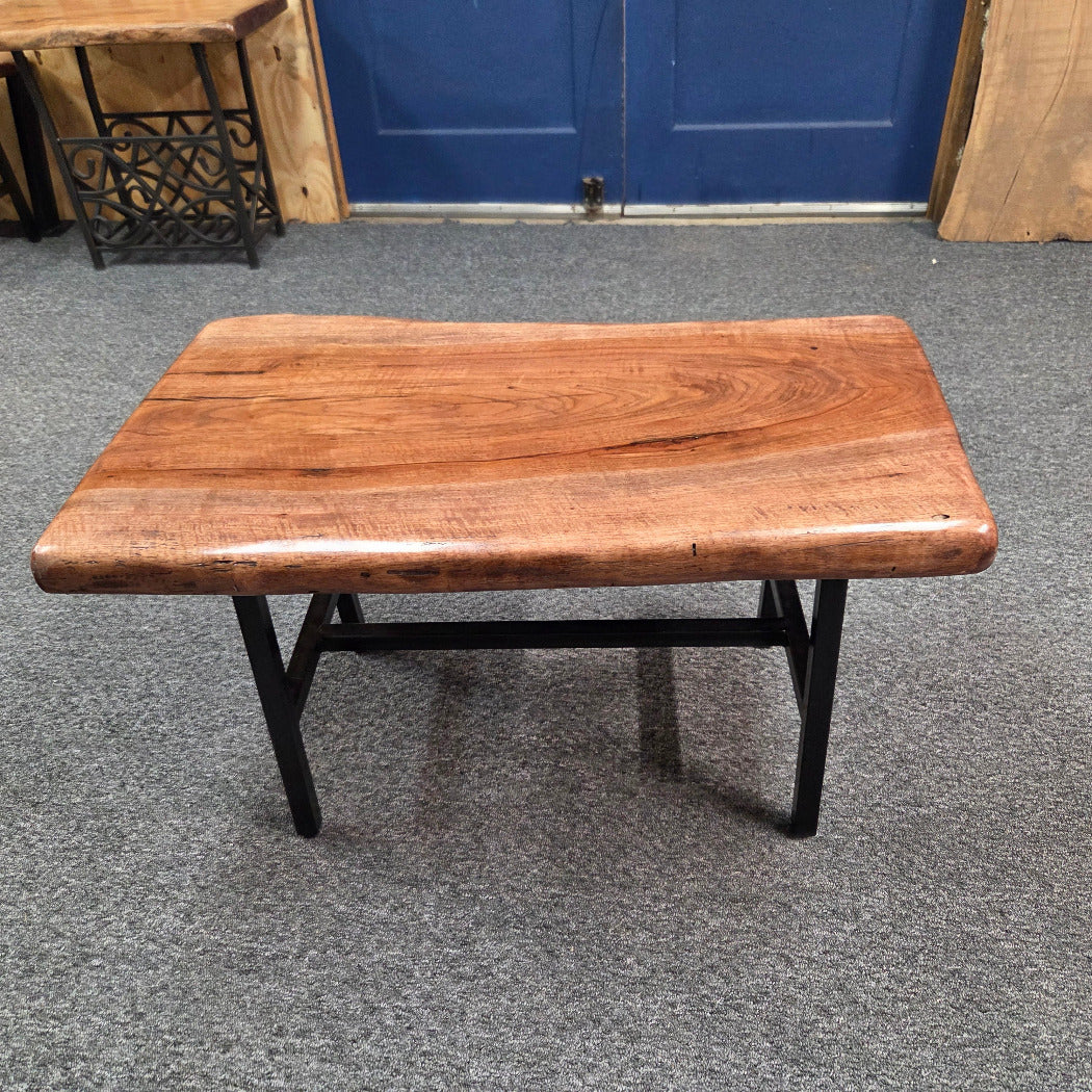 A rustic live-edge coffee table with a warm brown wooden top and a black metal frame, set on a gray carpeted floor in front of a blue door.