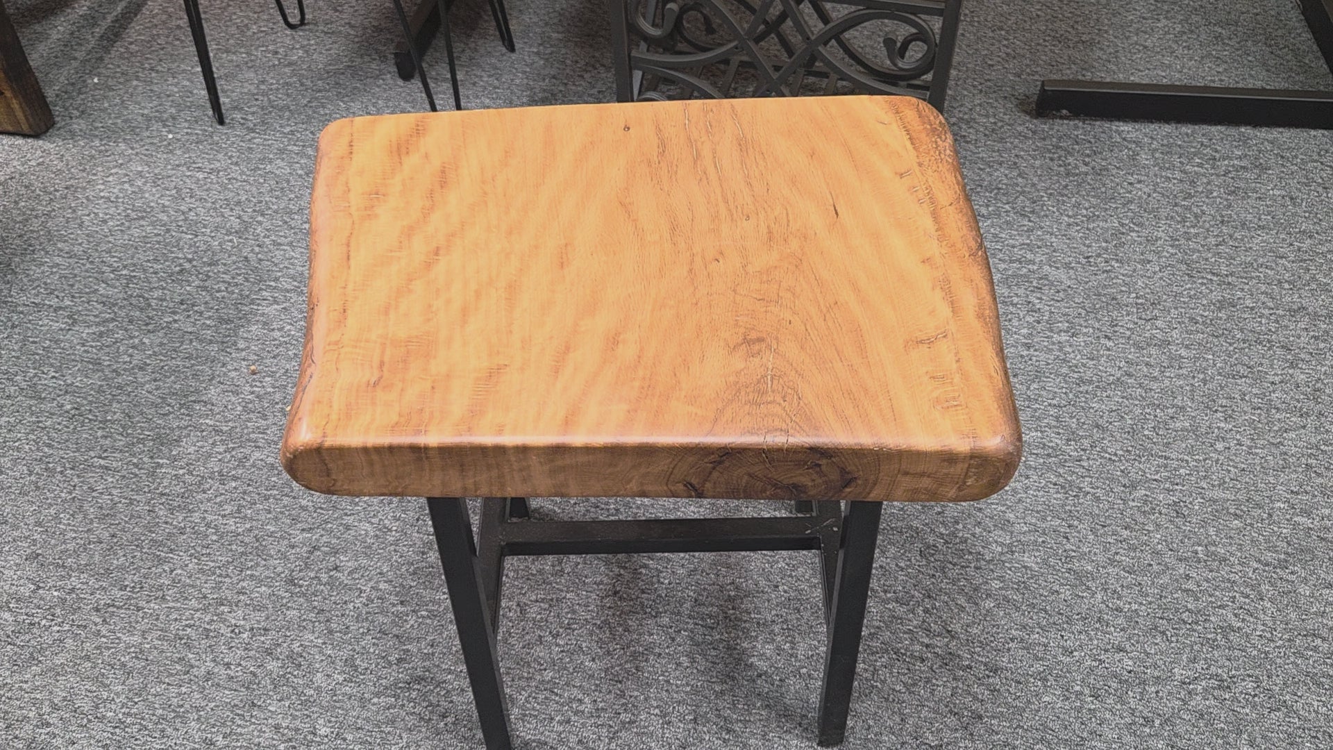 A rustic Live Oak end table with a thick, live-edge natural wood seat and a black metal frame, set on a gray carpeted floor.