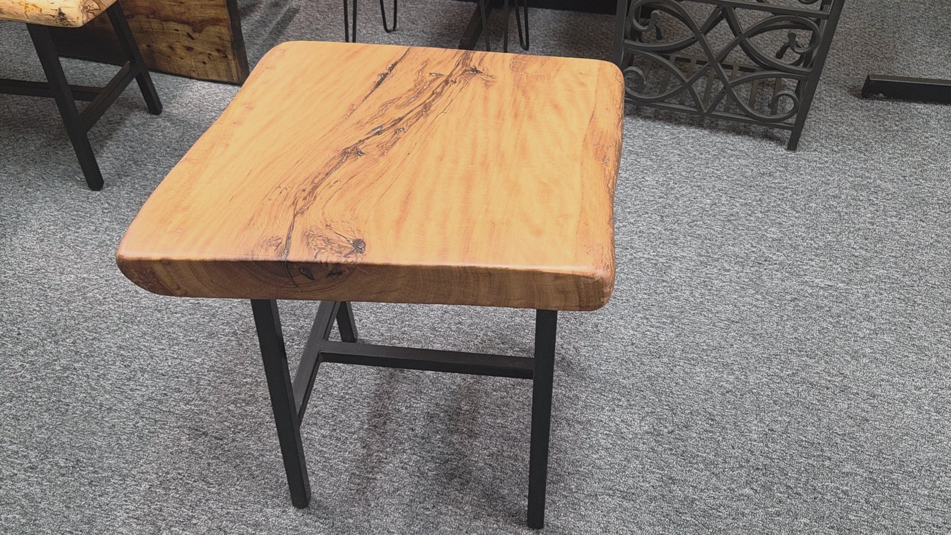 A rustic live-edge Live Oak end table with a square top and black metal legs, photographed on a gray carpeted floor.