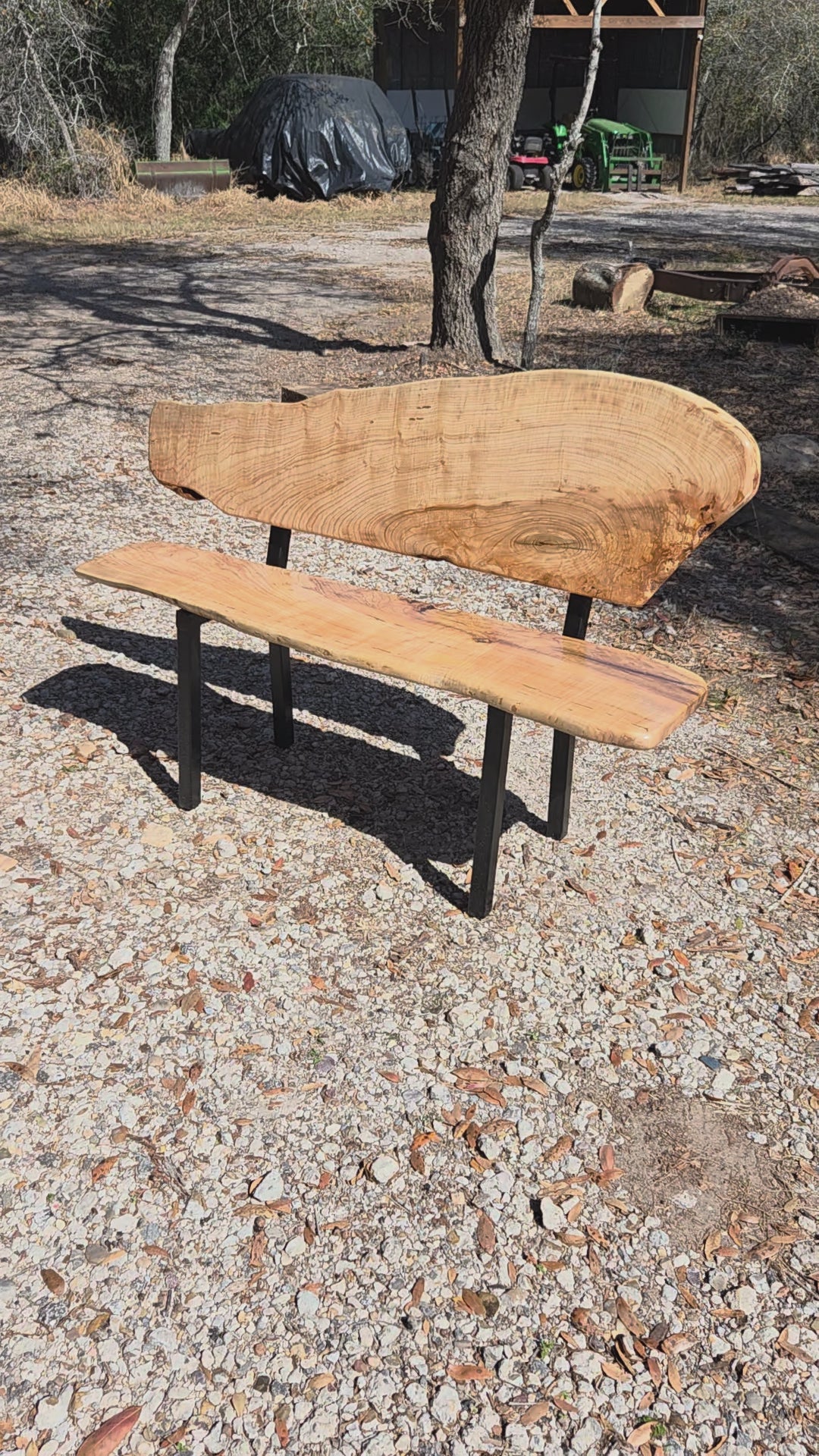 A rustic Ash live-edge wooden bench with a thick natural wood backrest and seat supported by black metal legs, photographed outdoors on gravel.