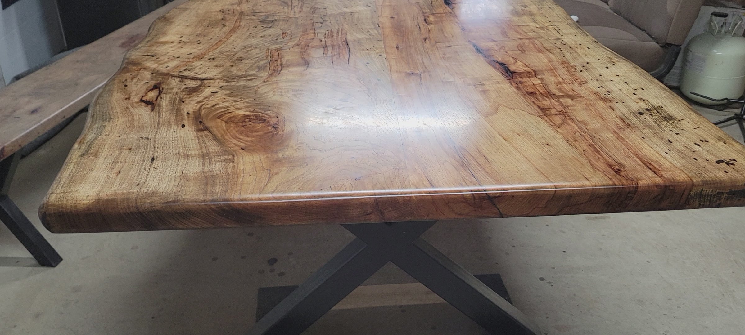 A rustic live-edge Pecan dining table with a natural wood top and black metal X-style legs, photographed in a workshop or warehouse setting.