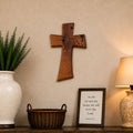 A warm, rustic interior scene featuring a wooden wall cross mounted above a matching wooden console table. The table is styled with a large cream ceramic vase containing green foliage, a small wicker basket, a framed scripture print, and a lit table lamp with a beige shade.