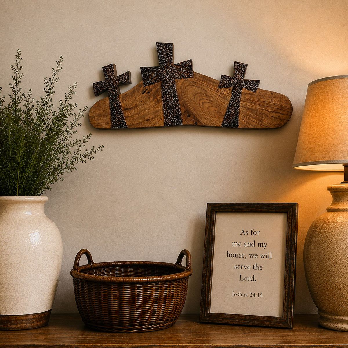 Hand carved wooden three cross wall art and rustic wooden console table styled in a warm home setting, featuring a white ceramic vase with green foliage, a brown wicker basket, a framed scripture print, and a lit table lamp against a beige wall.